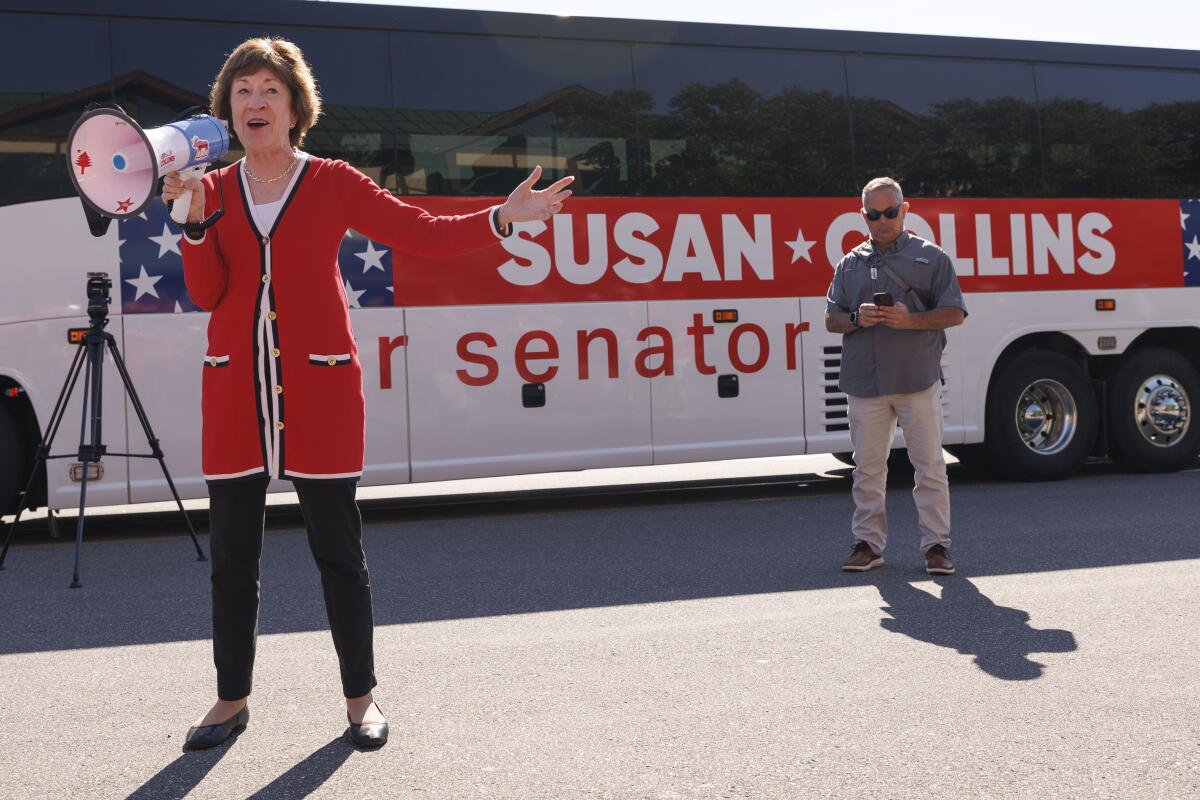 Susan Collins se paró frente a un autobús rojo y blanco con su nombre mientras sostenía un megáfono mientras los hombres miraban.
