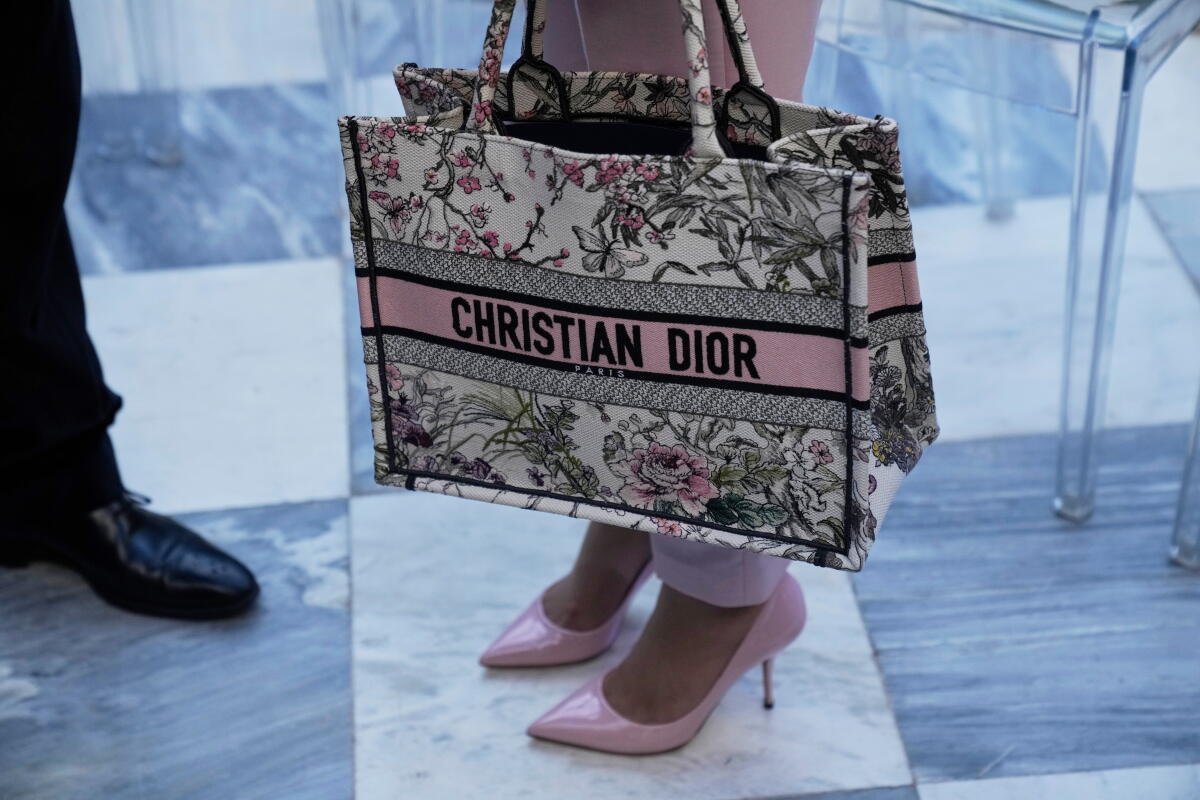 Una mujer viste pantalones rosas y tacones altos rosas y lleva un bolso de mano Christian Dior con un diseño floral. 